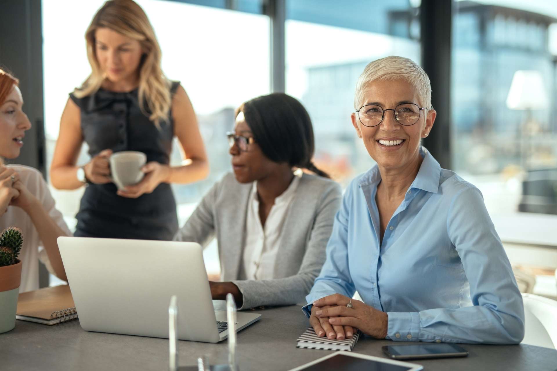 A group of professional women collaborating in a modern office, with one woman smiling at the camera while colleagues discuss work around a laptop during a team meeting.